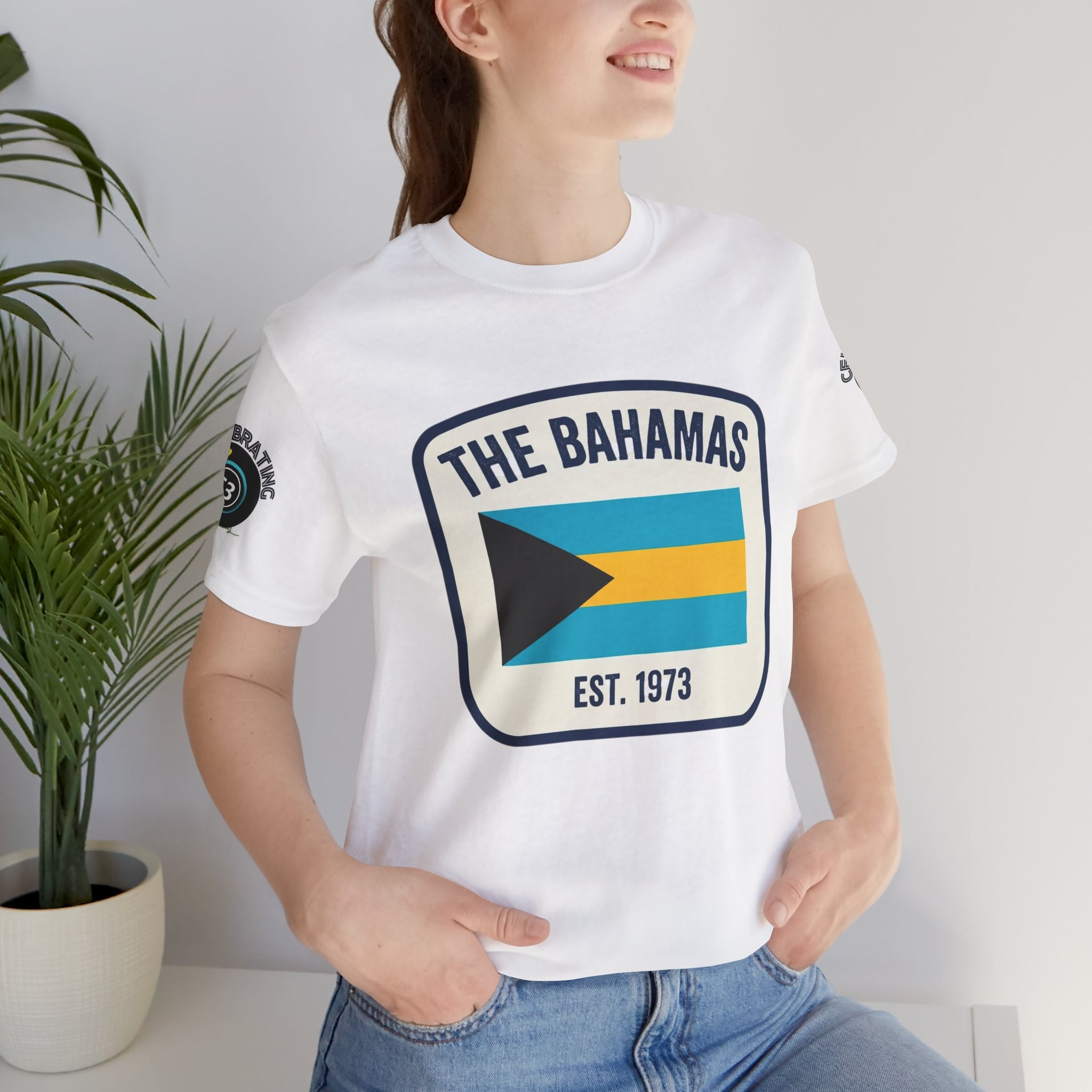 Person wearing a white t-shirt with 'The Bahamas' design, standing indoors next to a plant.
